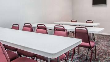 Un salón de conferencias con mesas blancas y sillas rojas, piso alfombrado y una televisión de pantalla plana en la pared.