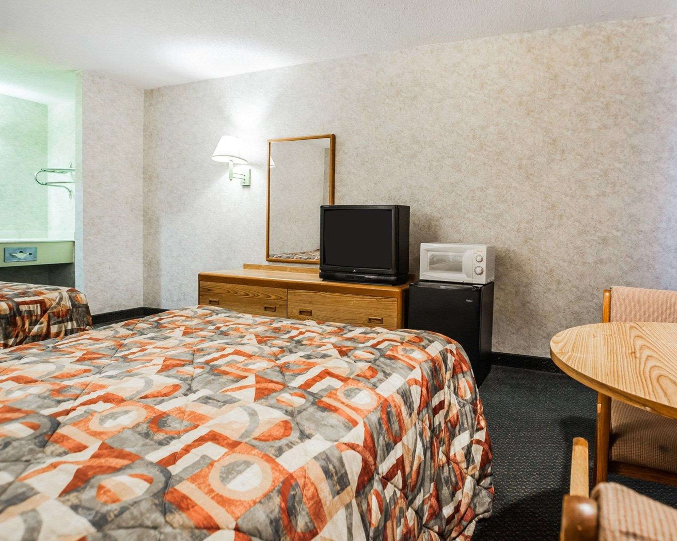 A hotel room with a bed, a small wooden dresser, a television, a microwave, a chair, and a lamp.