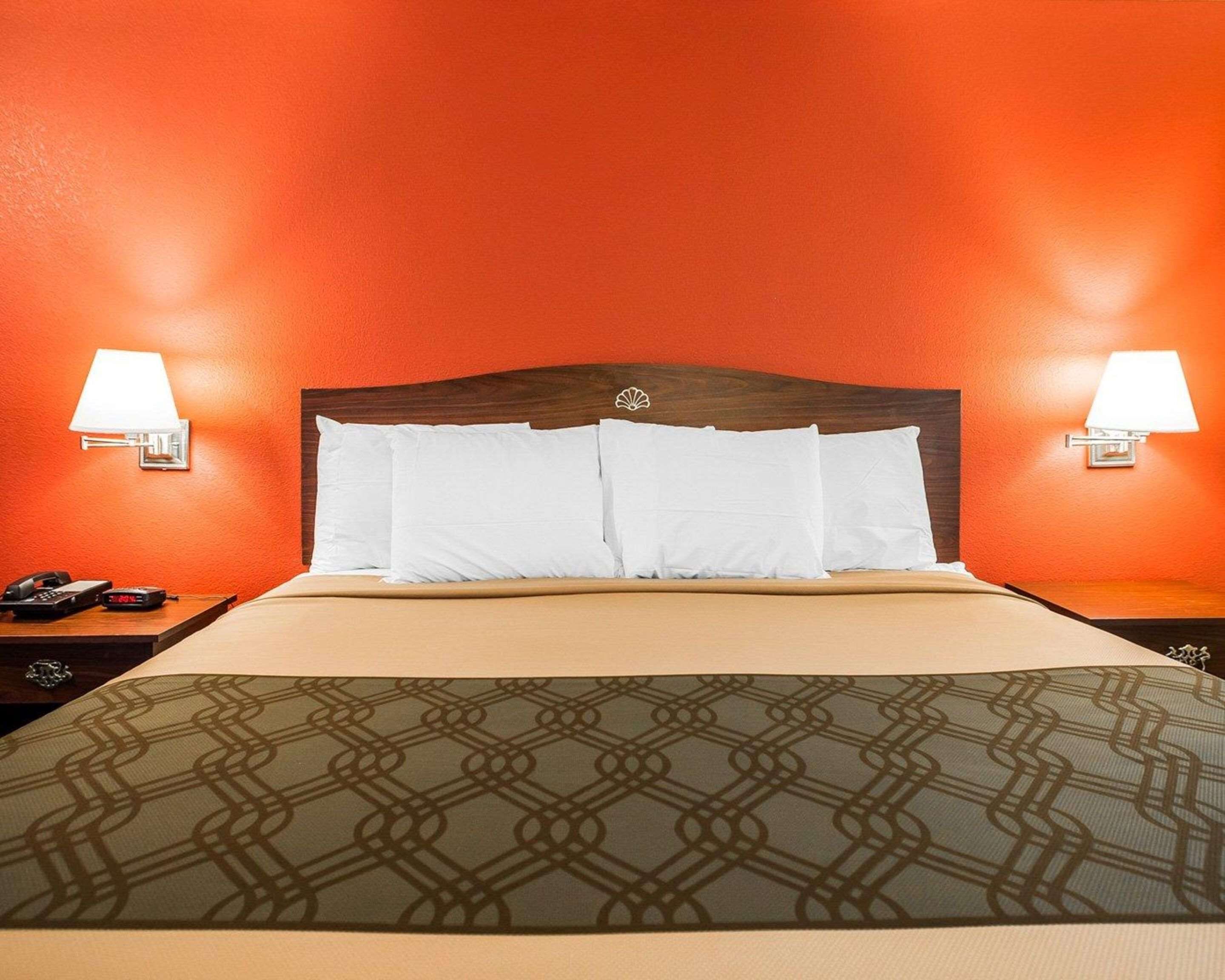 Una camera d'albergo con una parete arancione, una testiera in legno, cuscini bianchi, una coperta a motivi e due lampade ai comodini.