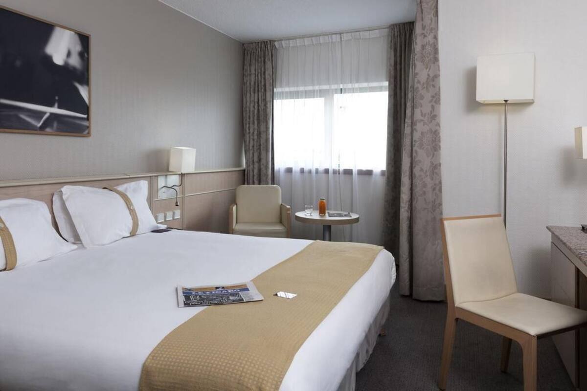 A hotel room with a large bed, a chair, a small table, a lamp, and a window with curtains.