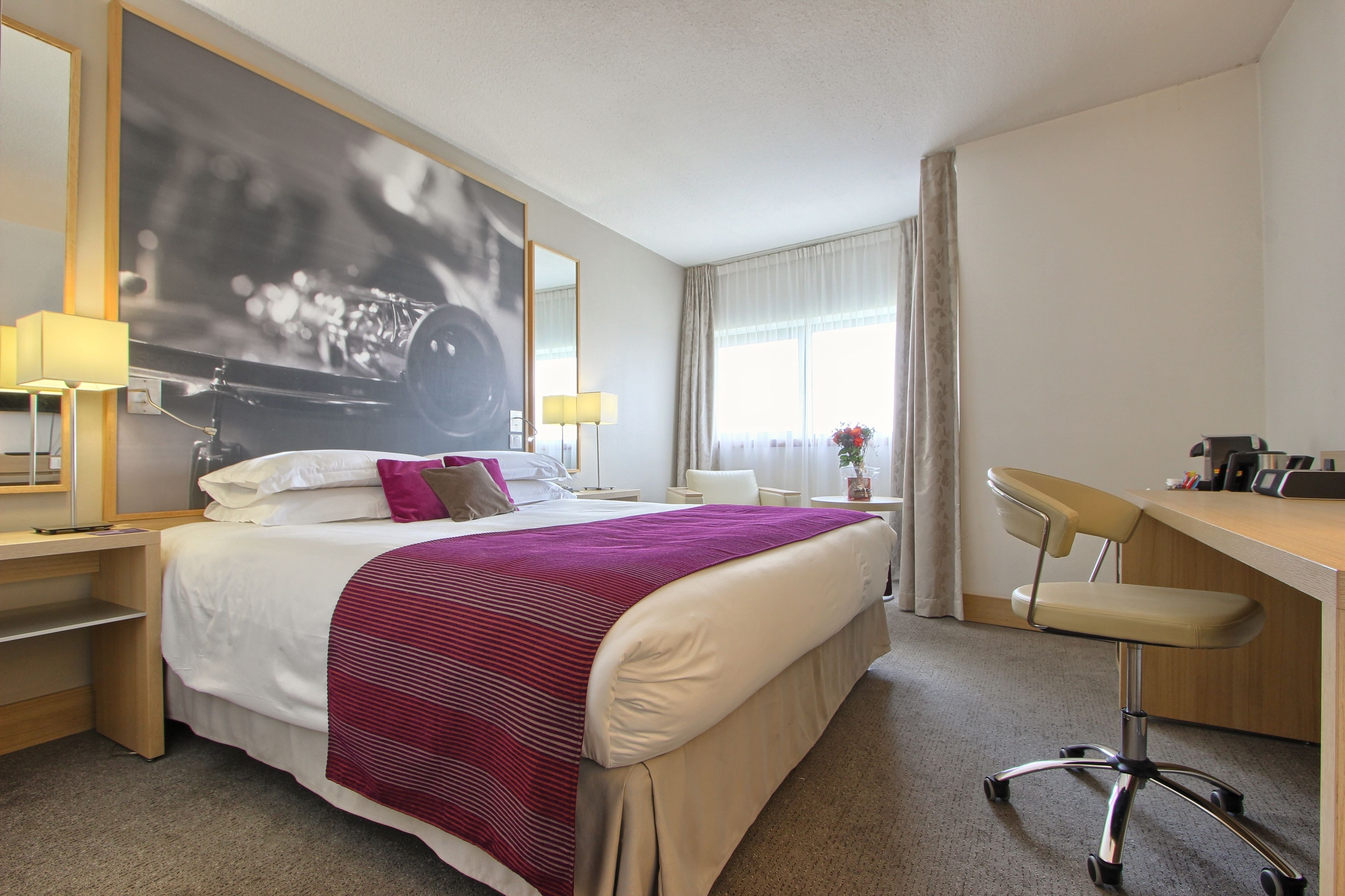 A hotel room with a large bed, a desk, a chair, a lamp, a mirror, and a window with curtains.