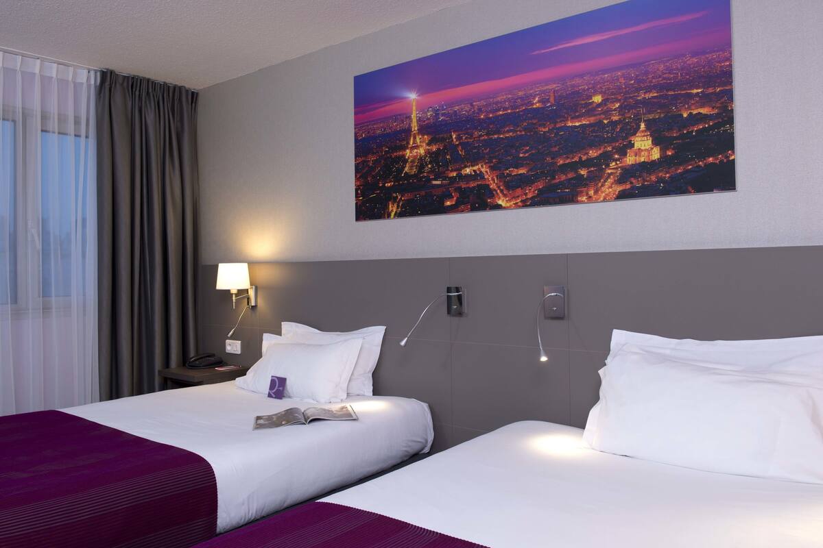 Une chambre d’hôtel avec deux lits, une table de chevet, une lampe, un tableau représentant un paysage urbain et une fenêtre avec des rideaux.