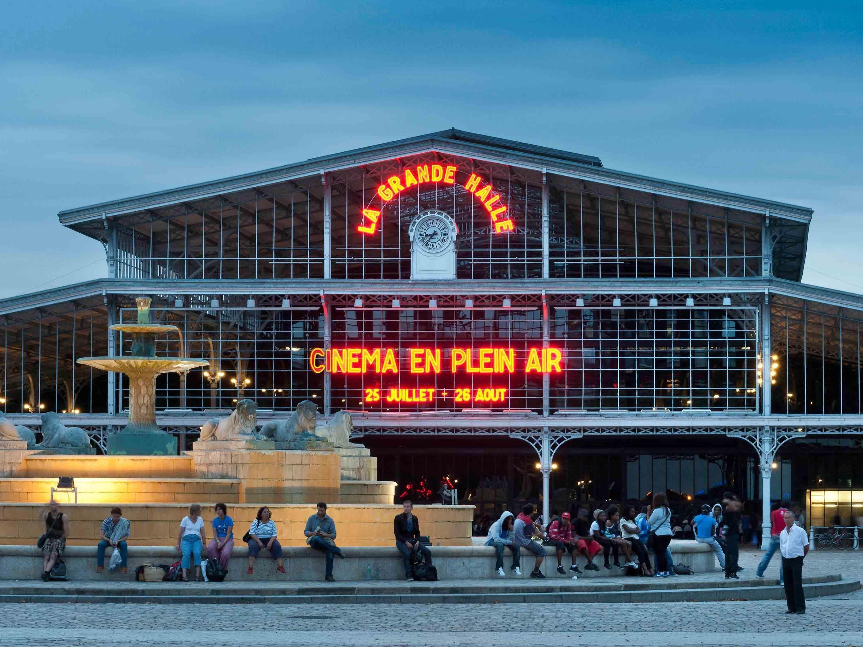 A large building with a prominent sign reading 'CINEMA EN PLEIN AIR' and a fountain in front.