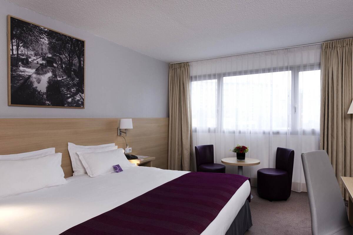 Une chambre d’hôtel avec un grand lit, un bureau, deux chaises, une petite table ornée d’un vase de fleurs et un tableau accroché au mur.