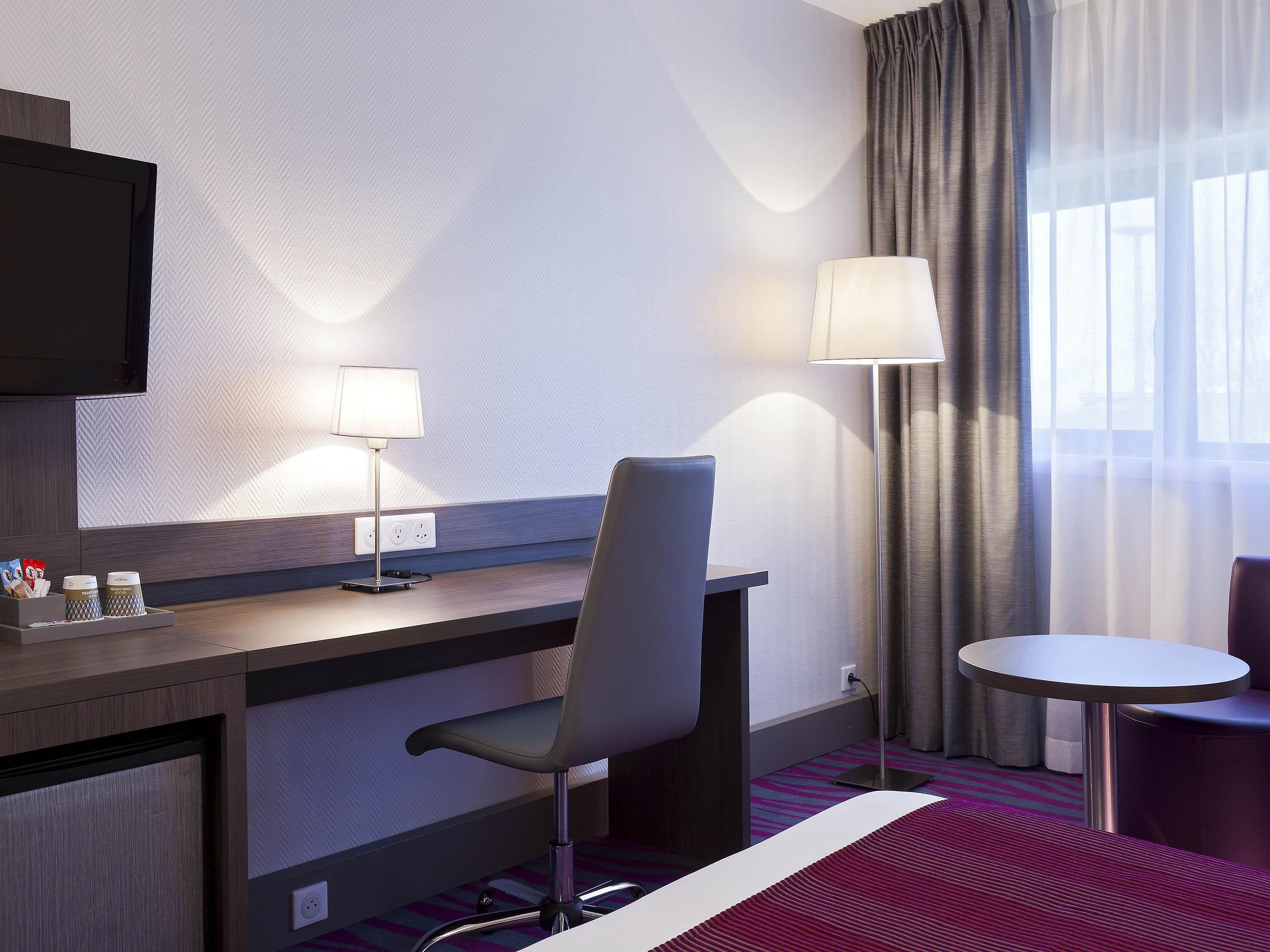 A hotel room with a desk, chair, TV, lamp, and a small round table.