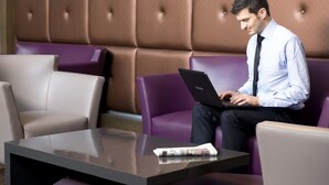Un homme est assis sur un canapé violet et utilise un ordinateur portable dans le hall d’un hôtel moderne. On y voit un panneau de tête de lit marron à motifs et une table basse en verre.