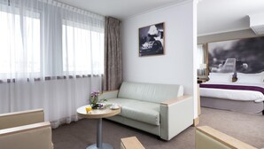 Une chambre d’hôtel avec un canapé vert clair, une table ronde sur laquelle se trouvent une boisson et des fleurs, un lit avec une literie blanche et une grande fenêtre ornée de rideaux légers.