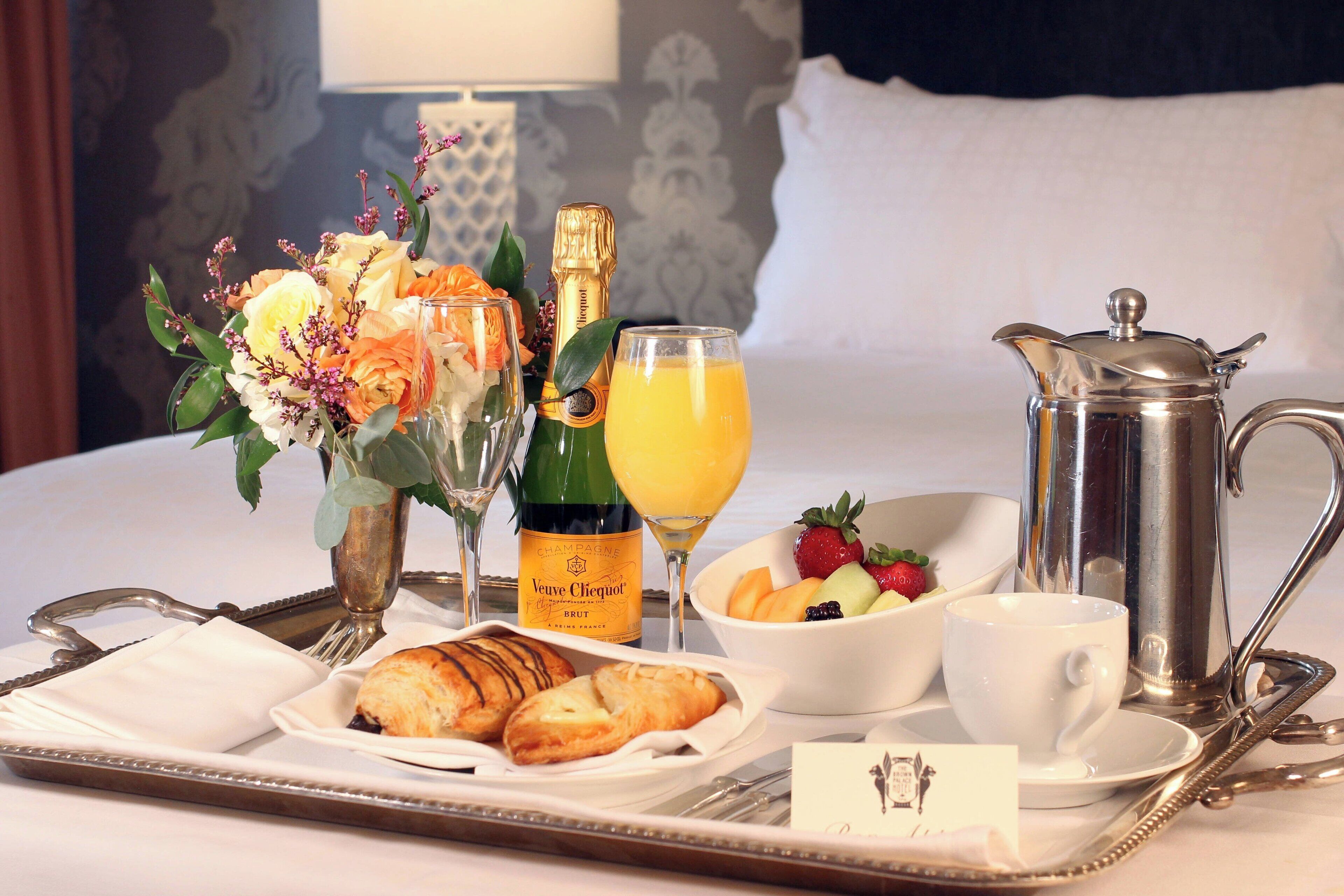 A breakfast tray with croissants, a bottle of Veuve Clicquot champagne, a glass of orange juice, a fruit bowl, a coffee pot, and a white cup on a hotel bed.
