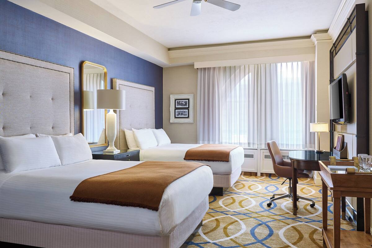 Habitación de hotel con dos camas, un escritorio, una silla, un televisor y un ventanal con cortinas.