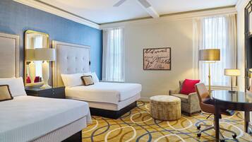 A hotel room with two beds, a ceiling fan, a round table with chairs, a small ottoman, and a large mirror.