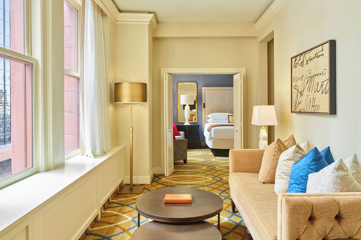 Habitación de hotel con un ventanal grande, una mesa de centro redonda, un sofá con cojines, una cama y un espejo.