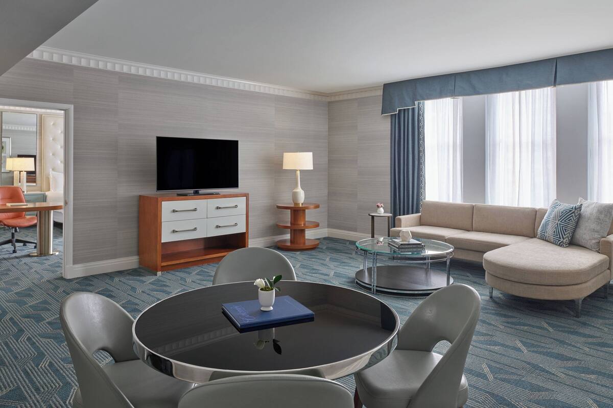 Una habitación de hotel con televisión de pantalla plana, un sofá, una mesa de comedor redonda con sillas, una pequeña mesa de centro y un amplio ventanal con cortinas.
