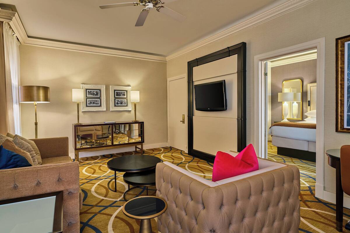 Una habitación de hotel con un sillón capitonado, un sofá, una mesita redonda, un televisor montado en la pared y una cama con una mesita de noche.