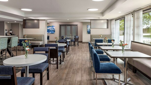 Un hall d’hôtel moderne avec des tables rondes, des chaises rembourrées en tissu bleu et de grandes fenêtres donnant sur l’extérieur.