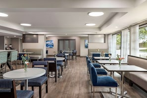 Un moderno vestíbulo de hotel con mesas redondas, sillas tapizadas en azul y grandes ventanales con vistas al exterior.