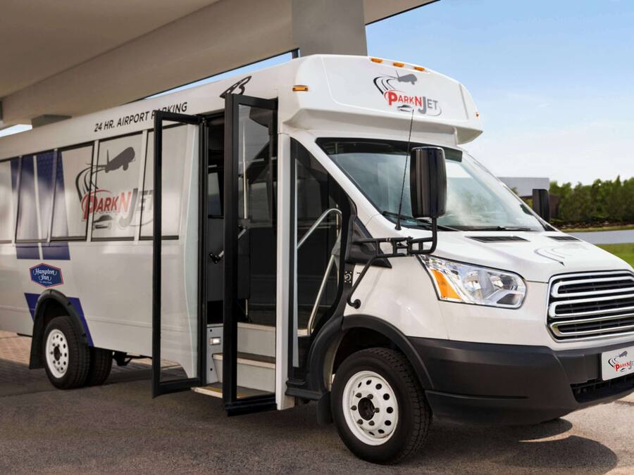 A white bus with a side door open, displaying a "24 HR. AIRPORT PARKING" sign and "Park N Jet" branding.
