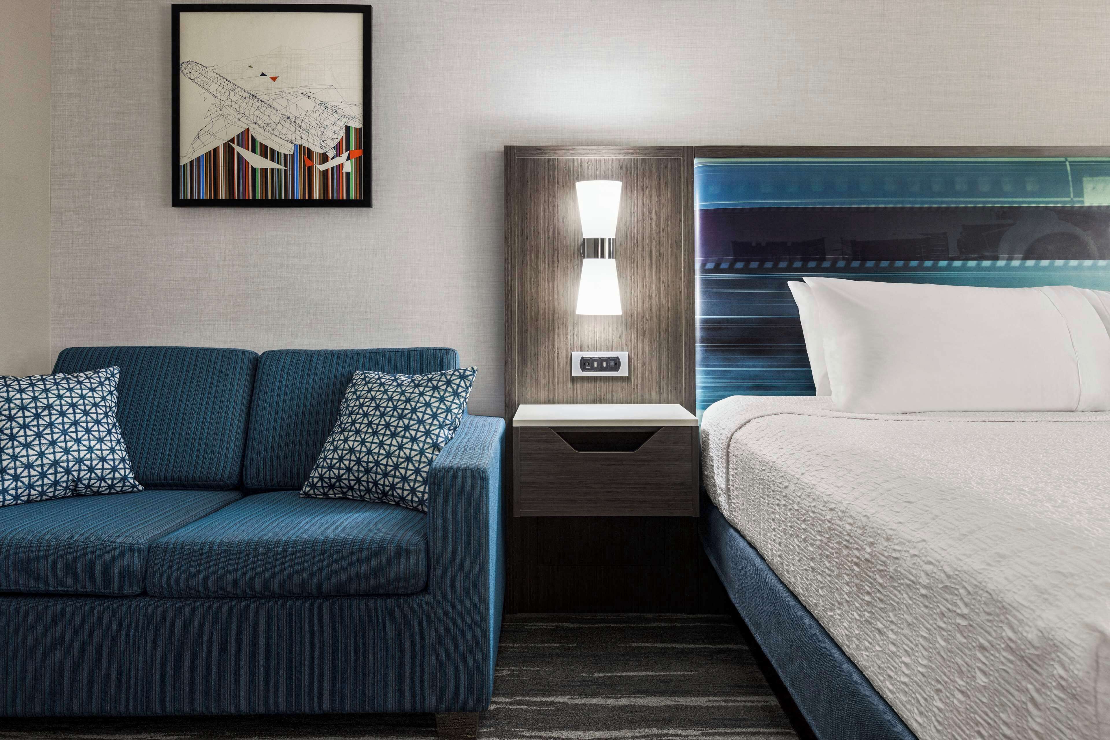 A hotel room with a blue sofa, a bed with white bedding, a nightstand with a lamp, and a framed picture on the wall.