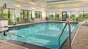 Une piscine intérieure avec de grandes baies vitrées, des chaises vertes et une terrasse carrelée.