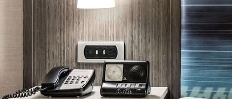 A hotel room with a wooden wall, a bedside table with a telephone and a power strip, and a window with blinds.