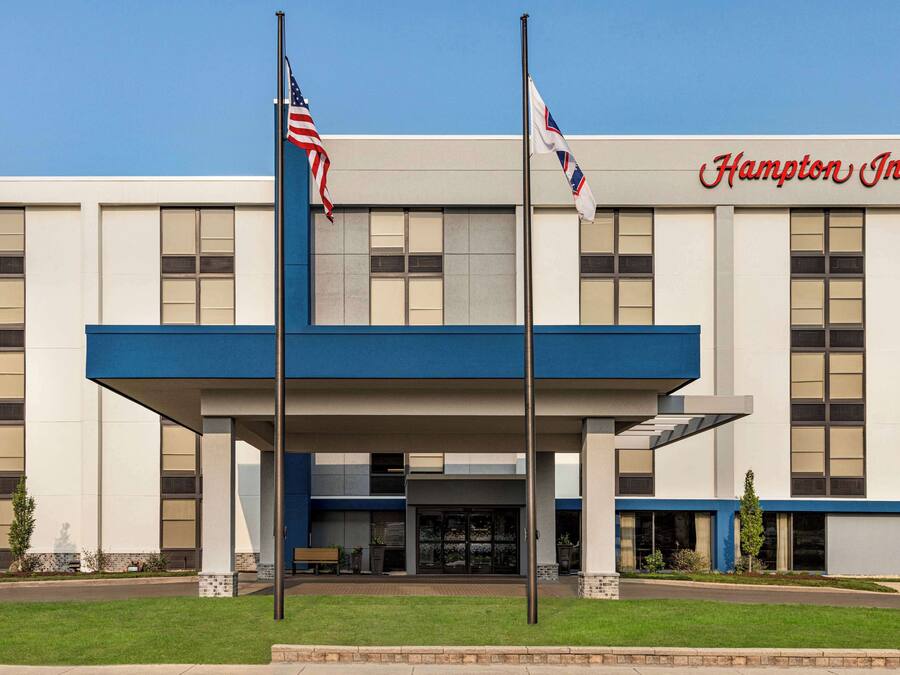 Un hotel Hampton Inn con fachada moderna, dos banderas y cielo despejado.