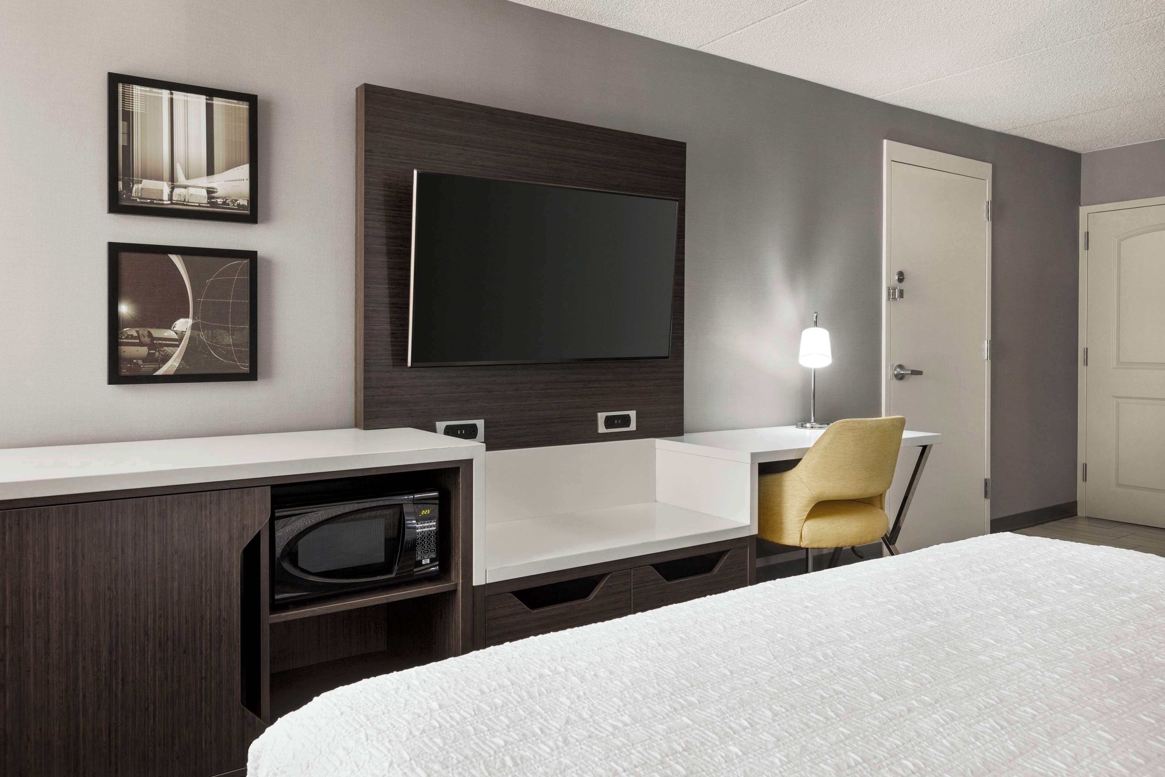 A hotel room with a bed, a desk with a chair, a flat-screen TV mounted on the wall, a microwave, and a door.