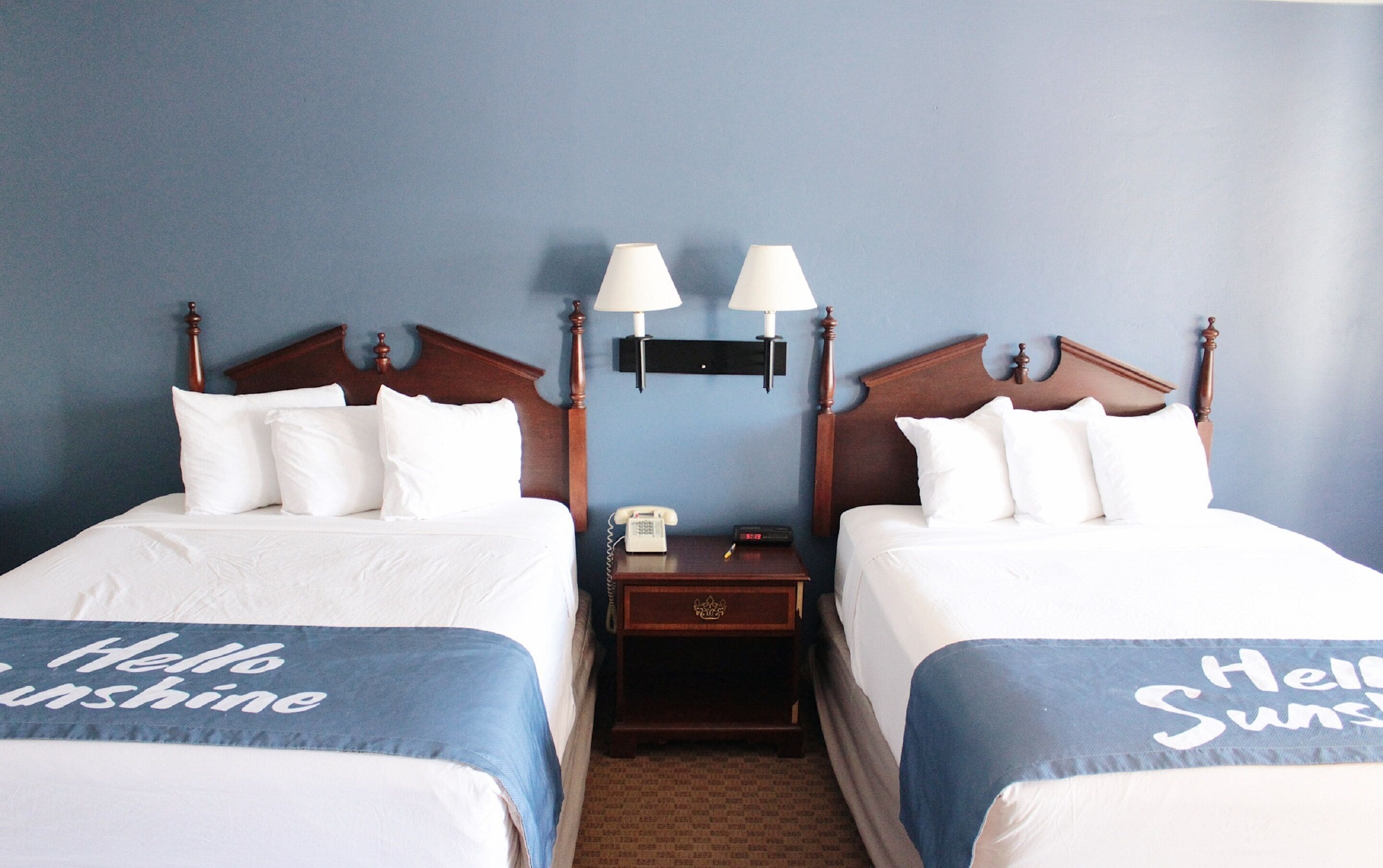 Habitación de hotel con dos camas, cada una con una colcha que dice 'Hello Sunshine', una mesita de noche con teléfono y reloj, y dos lámparas fijadas a la pared.