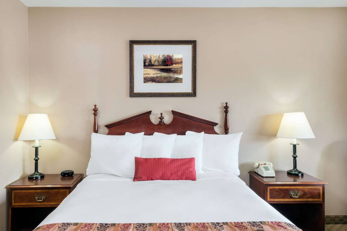 A neatly made bed with white and red pillows, a wooden headboard, and a framed picture on the wall. There are two bedside tables with lamps and a telephone on each.