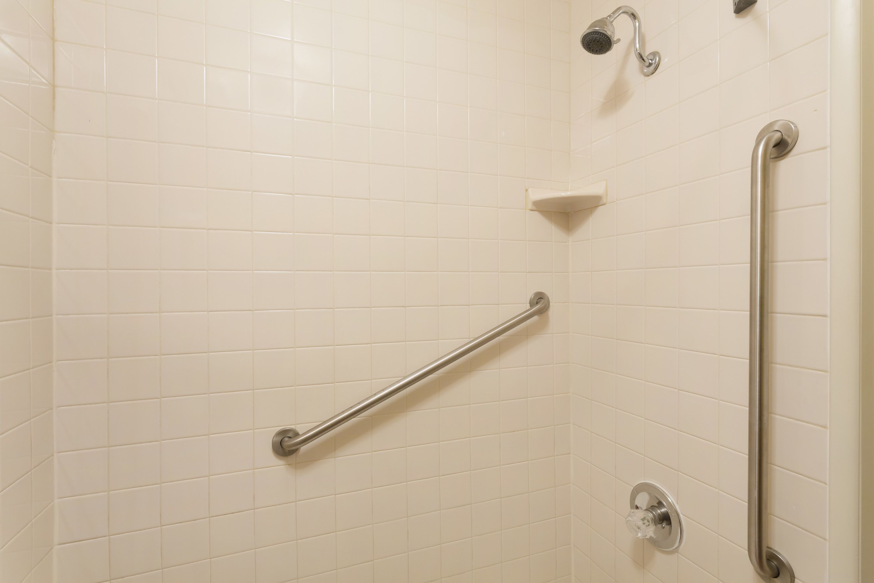 A bathroom with white tiled walls, a handheld showerhead, a grab bar, and a corner shelf.