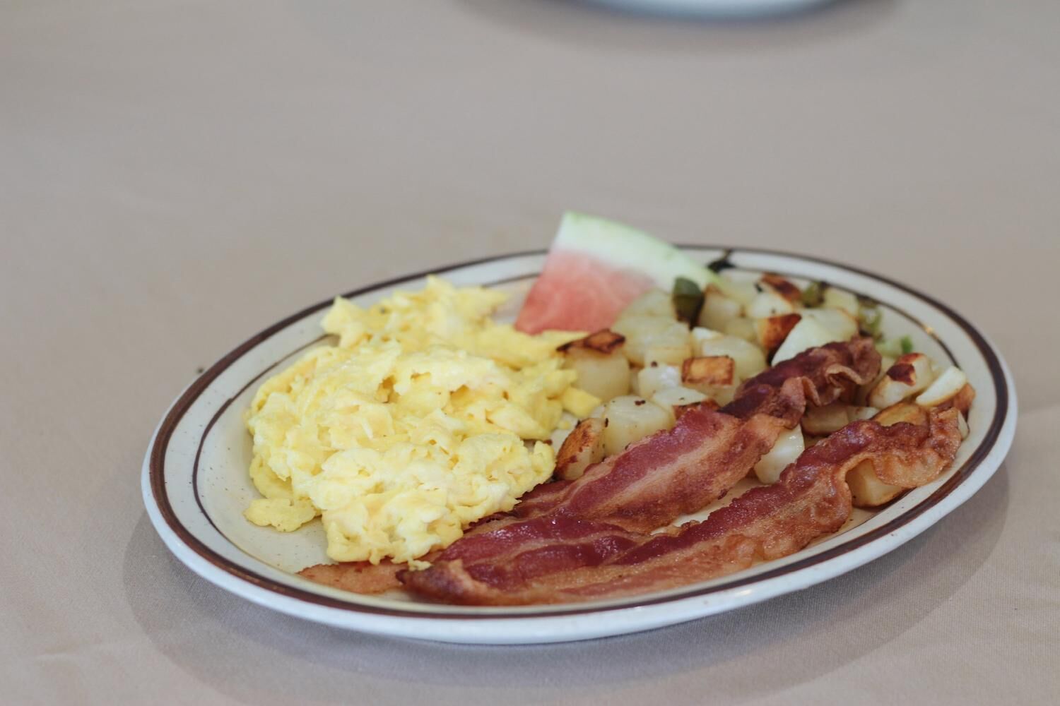 A plate of scrambled eggs, bacon, and diced potatoes with a slice of watermelon.