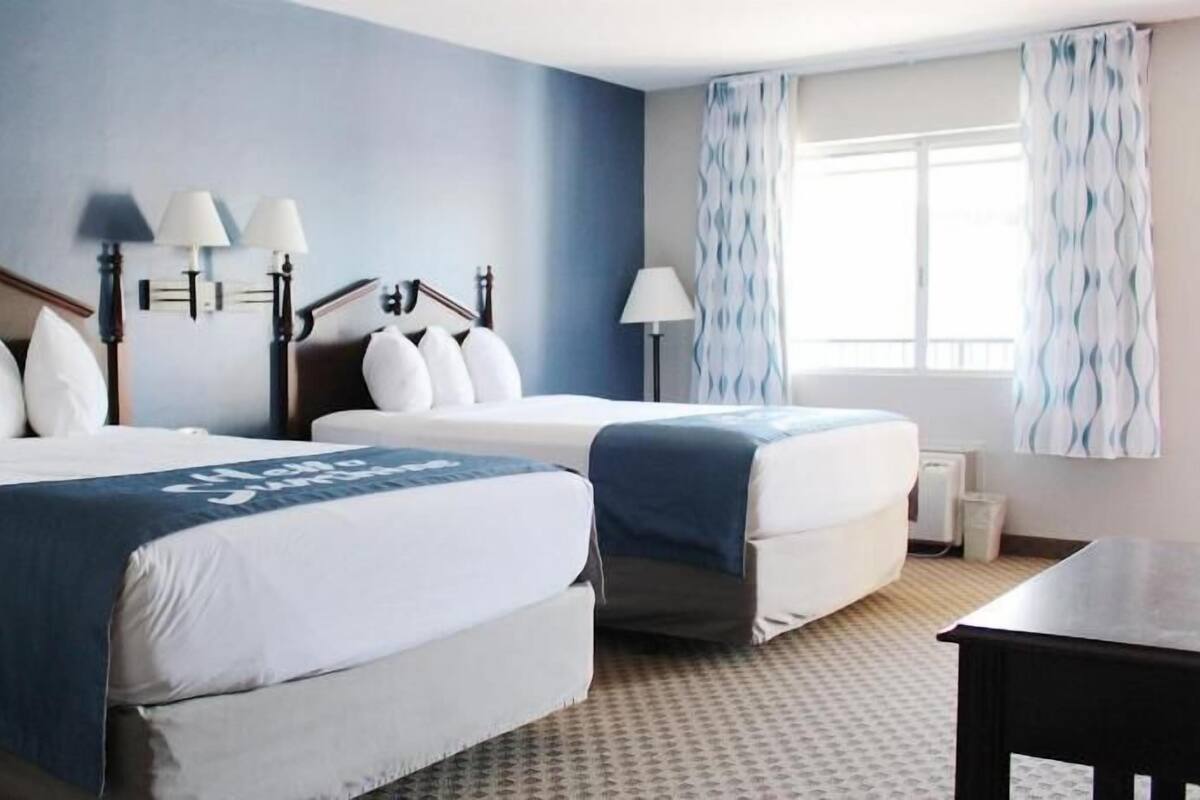 A hotel room with two beds, a desk, a window with curtains, and wall-mounted lamps.
