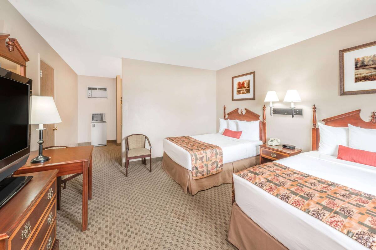 A hotel room with two beds, a desk with a television, a chair, and a small table with lamps.