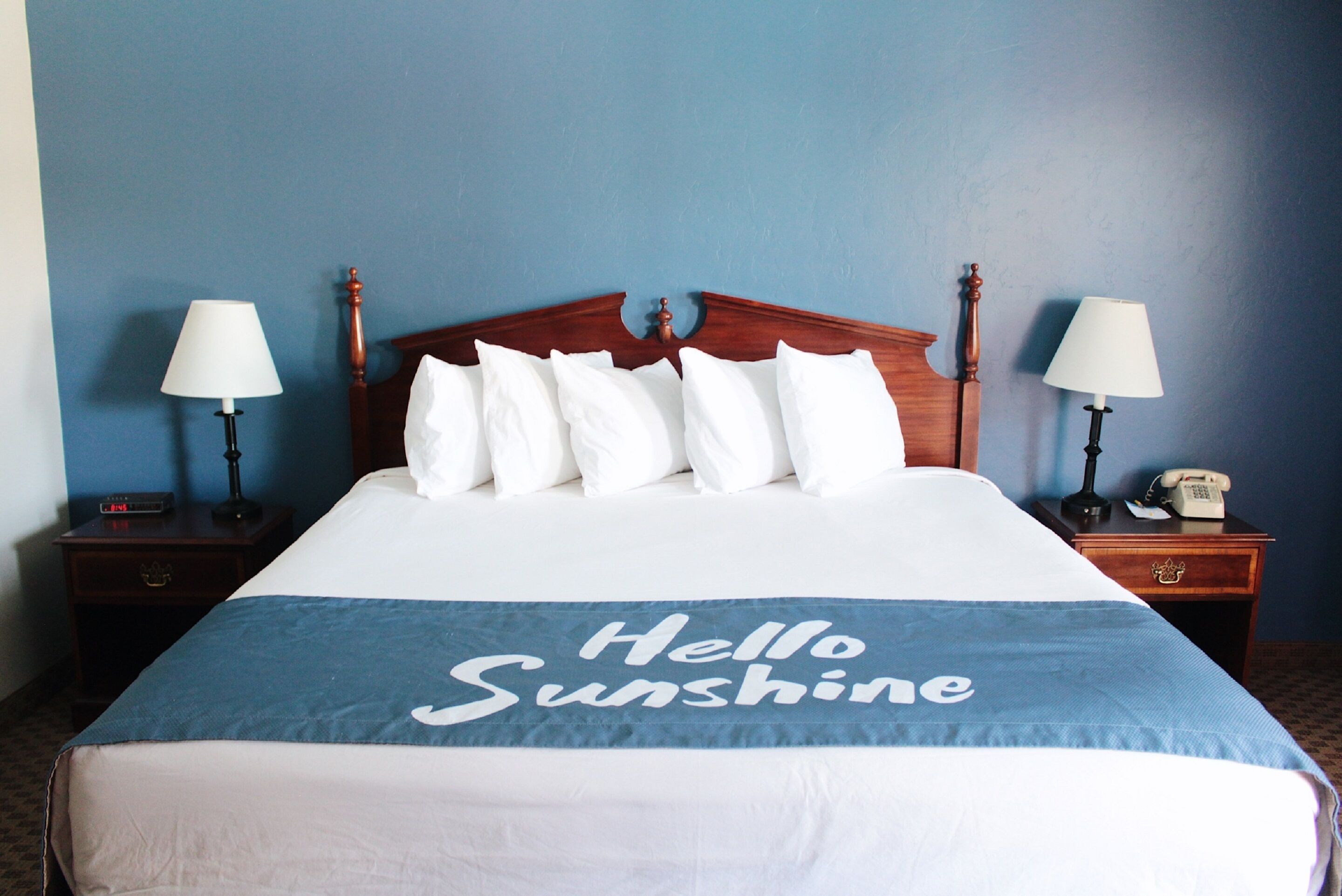 Una cama bien hecha con una colcha azul que dice “Hello Sunshine”, dos lámparas de noche, una mesita con un teléfono y un cabecero de madera.