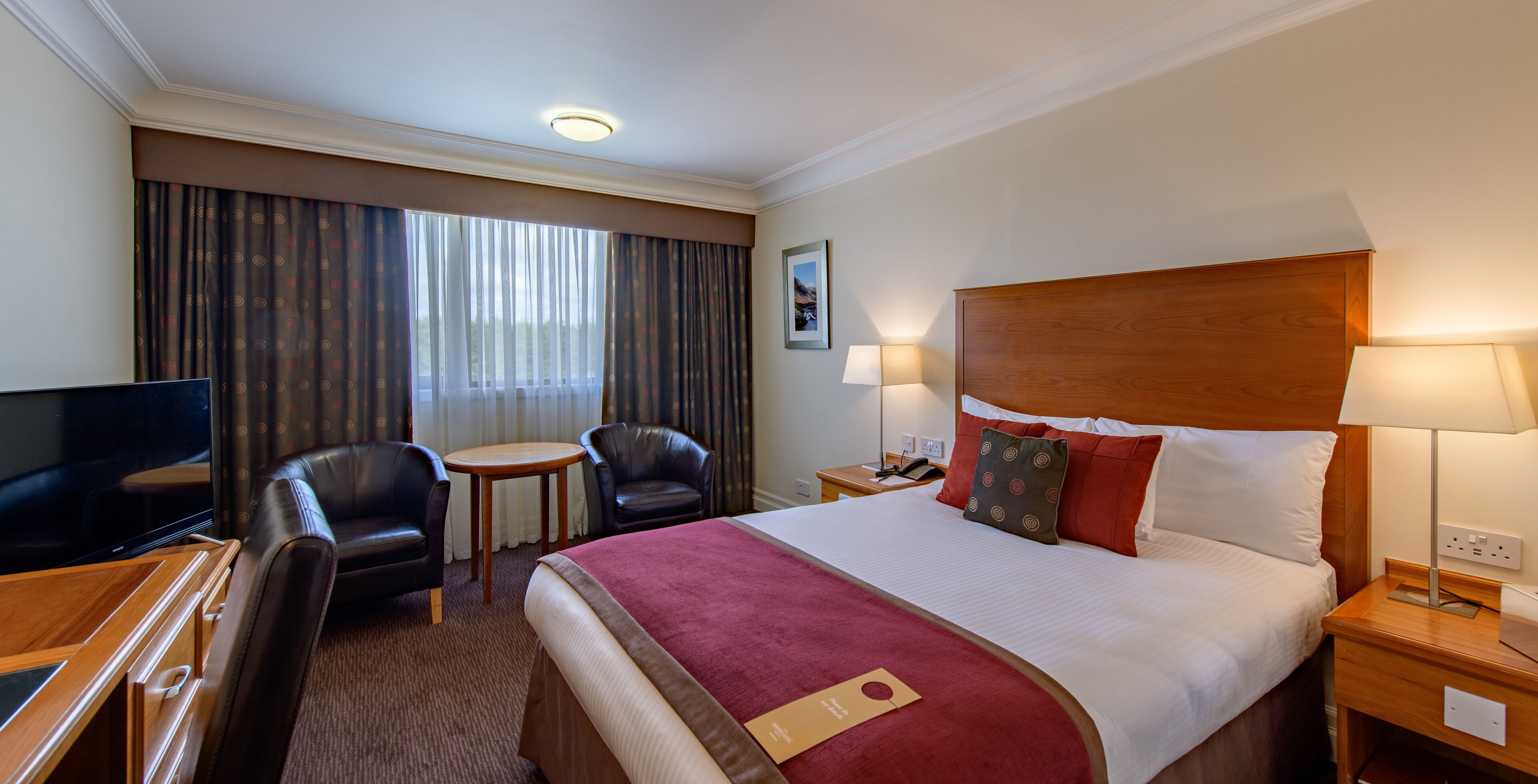 Quarto de hotel com cama, duas cadeiras, escrivaninha com televisão, mesa de cabeceira com abajur e janela com cortinas.