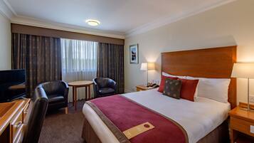 Quarto de hotel com cama, duas cadeiras, escrivaninha com televisão, mesa de cabeceira com abajur e janela com cortinas.