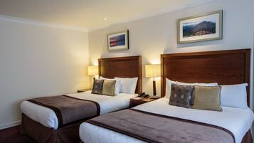 A hotel room with two beds, each with a wooden headboard, white bedding, and brown accents. There are two bedside lamps, a framed picture on the wall, and a small table with a phone.