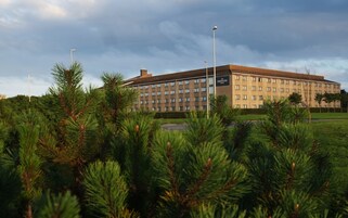 A large, multi-story hotel with a flat roof and a uniform facade, surrounded by a well-maintained lawn and pine trees.