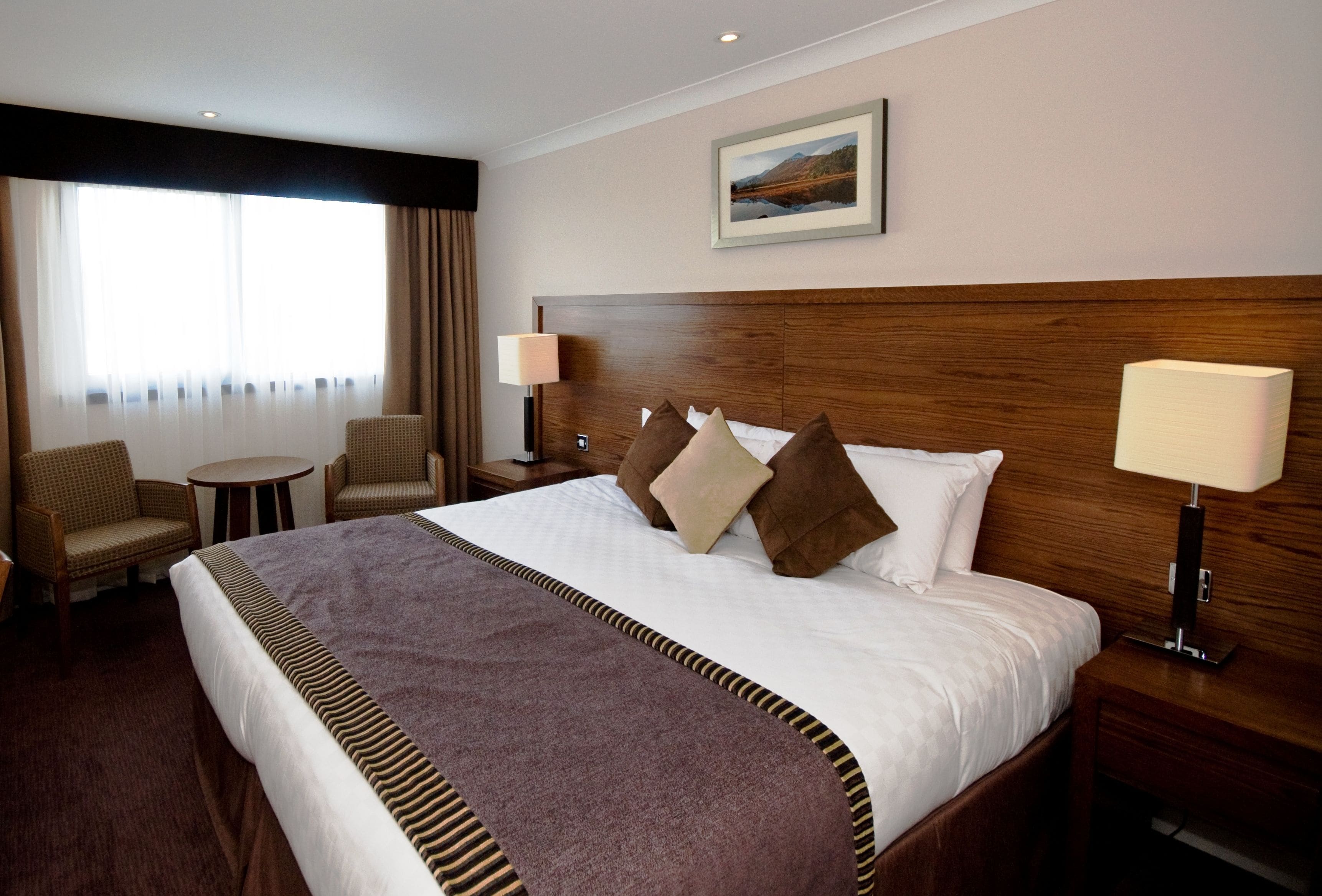 A hotel room with a large bed, two bedside tables, a nightstand, a chair, a round table, and a window with curtains.