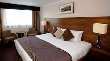 A hotel room with a large bed, two bedside tables, a nightstand, a chair, a round table, and a window with curtains.