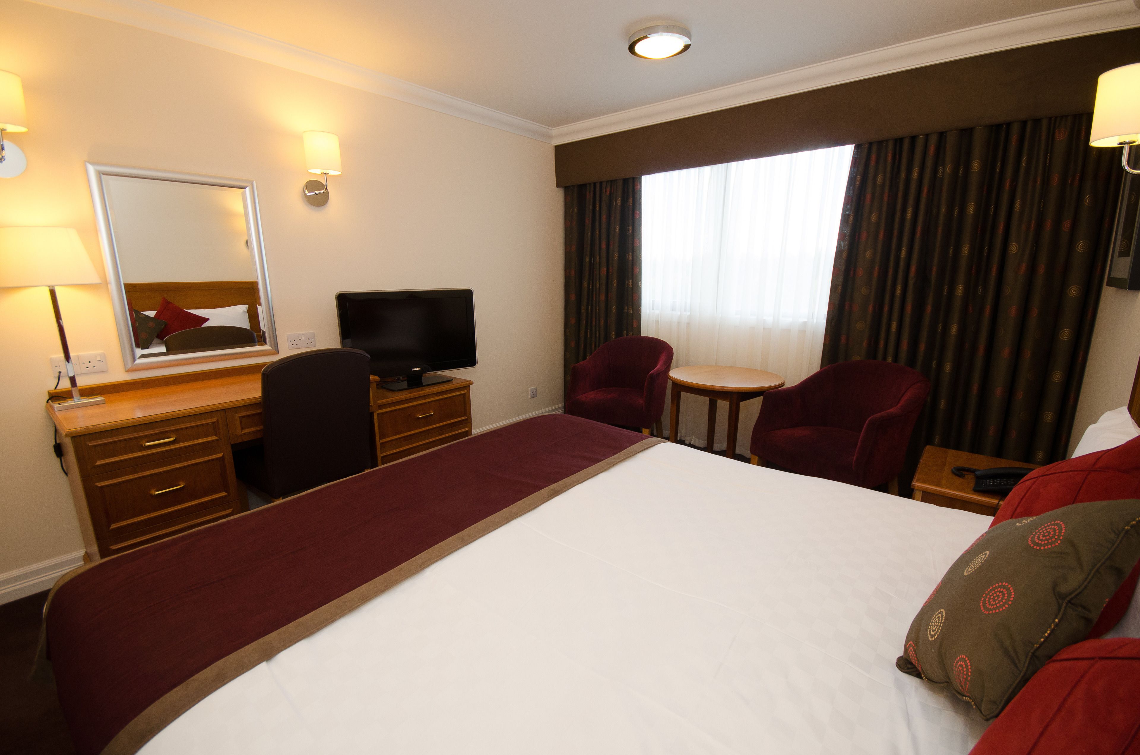 A hotel room with a large bed, a desk with a computer, two chairs, a TV, and a window with curtains.