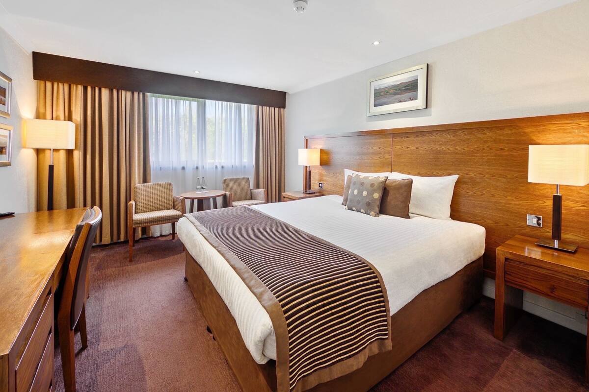 A hotel room with a large bed, a wooden desk, a chair, a small table, and two lamps.
