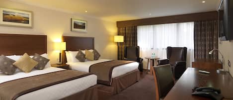 A hotel room with two beds, a desk with a telephone, a chair, a small table with a lamp, and a TV on the wall.