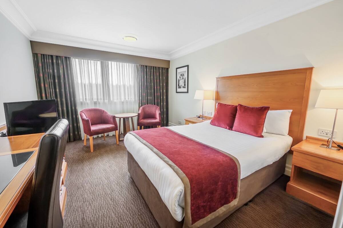 A hotel room with a large bed, a desk with a TV, two chairs, a wooden headboard, and a window with curtains.