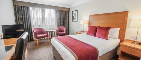 Habitación de hotel con una cama grande, un escritorio con televisor, dos sillas, cabecera de madera y una ventana con cortinas.