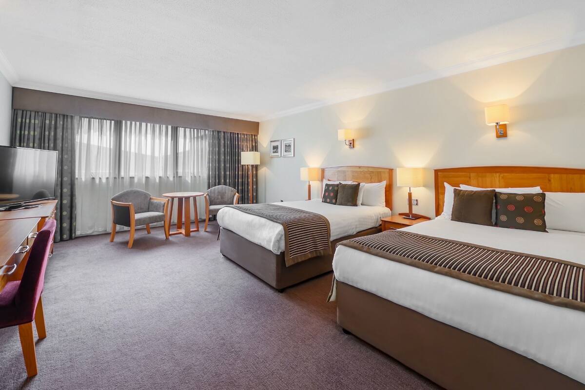 A hotel room with two beds, a desk with a chair, a TV, and a large window with curtains.
