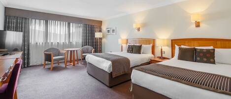 A hotel room with two beds, a desk with a chair, a TV, and a large window with curtains.
