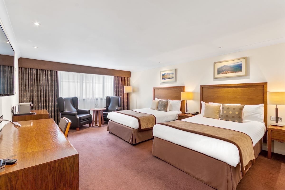 A hotel room with two beds, a desk, a chair, a small table, and a painting on the wall.