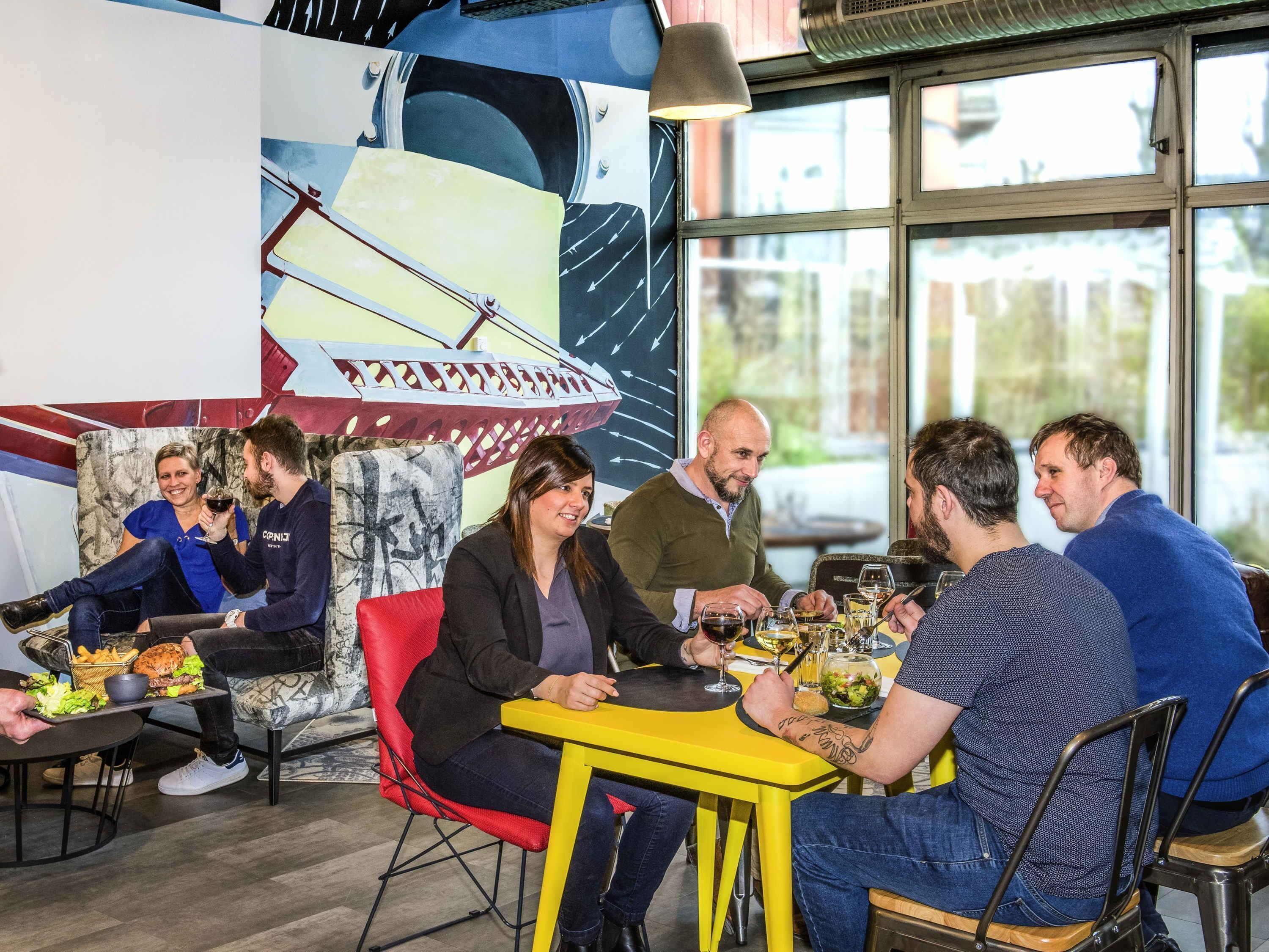 Eine Gruppe von Menschen speist in einem Restaurant mit modernem Interieur, inklusive eines großen, abstrakten Gemäldes an der Wand und großen Fenstern.