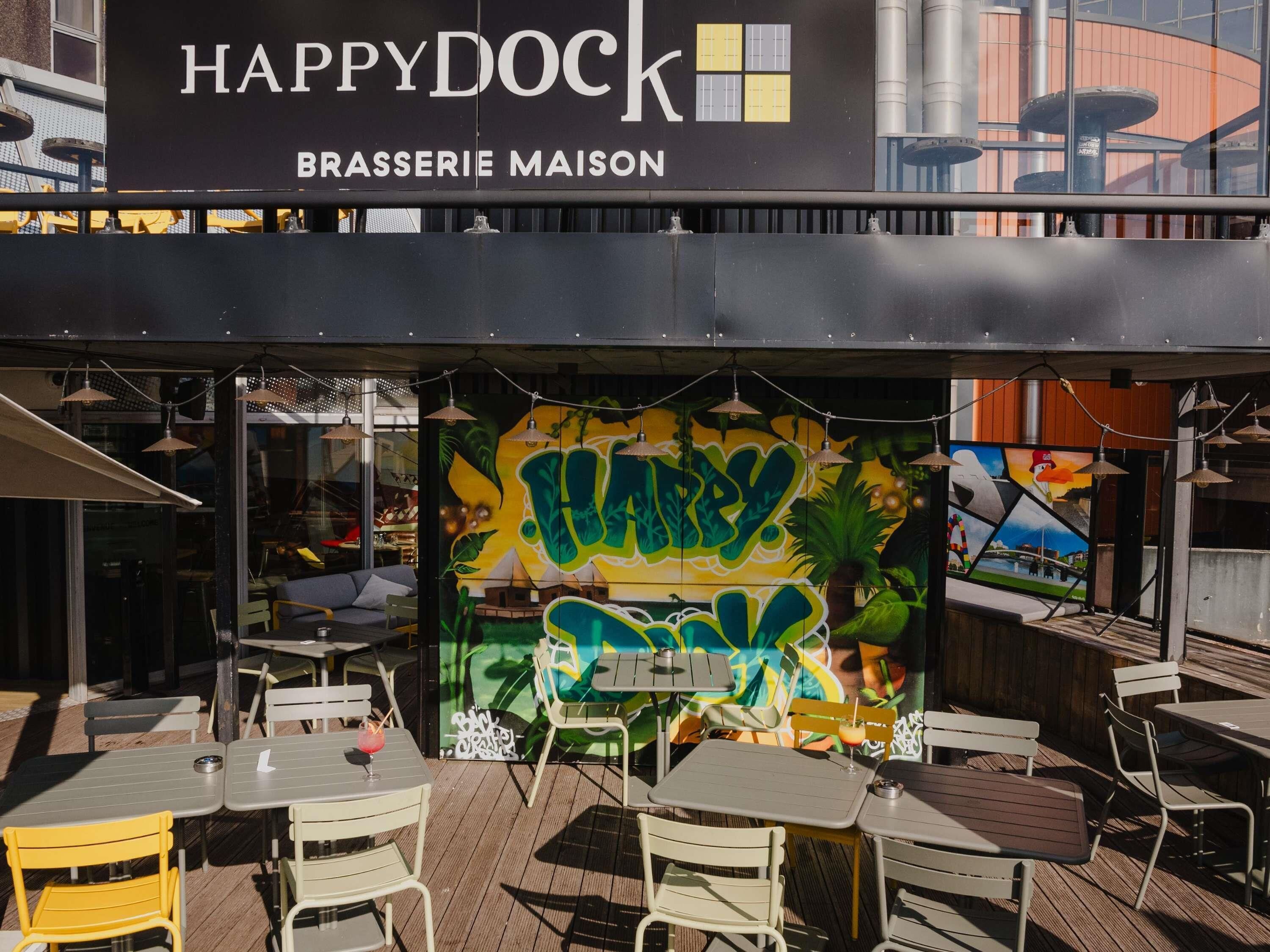 Un espace extérieur aménagé avec des tables et des chaises, une fresque murale ornée de graffitis et une enseigne indiquant : « HAPPY DOCK BRASSERIE MAISON ».