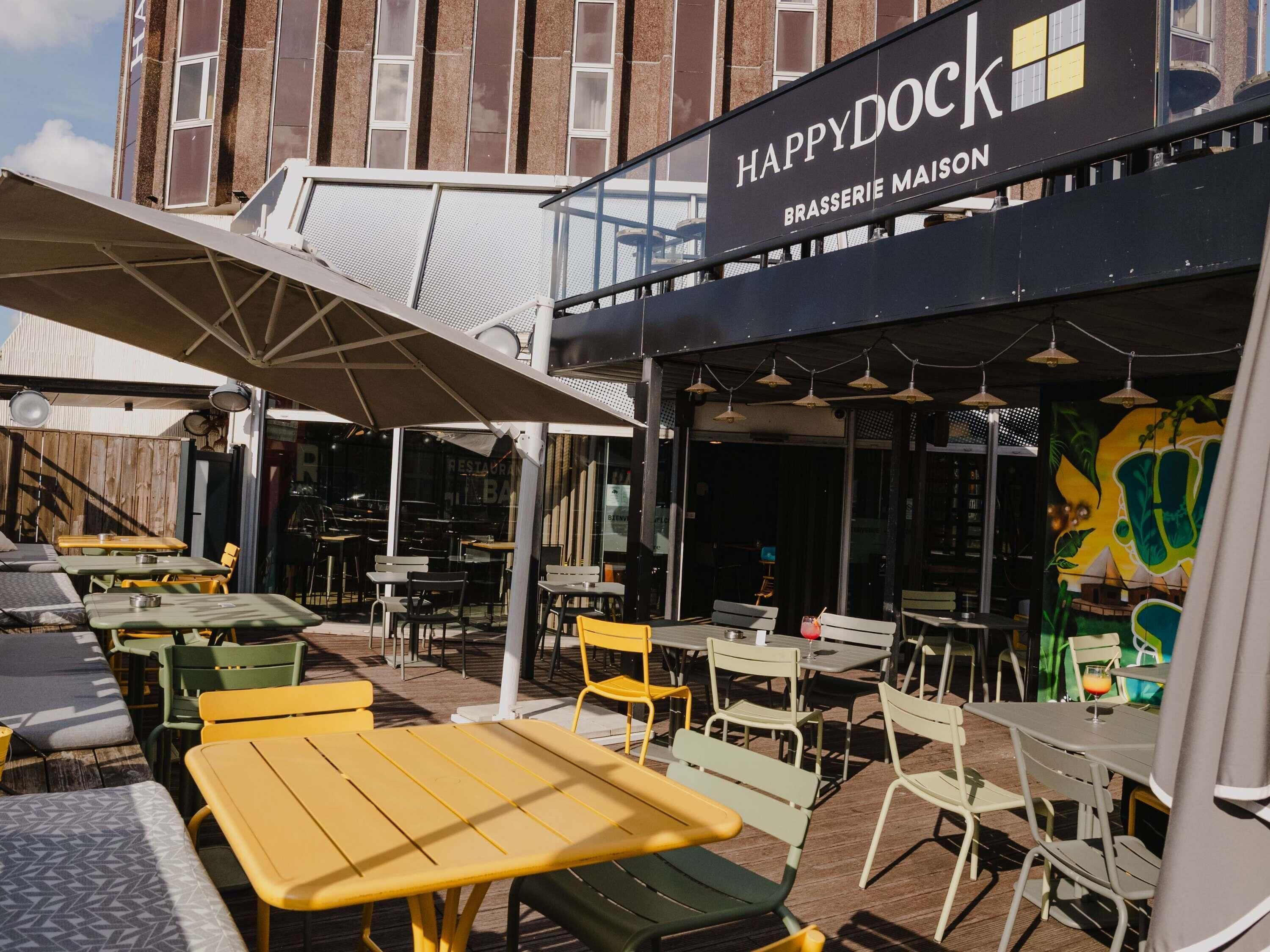 An outdoor seating area with tables and chairs, a covered section with a sign reading 'HAPPY DOCK BRASSERIE MAISON,' and a building with a brick facade in the background.