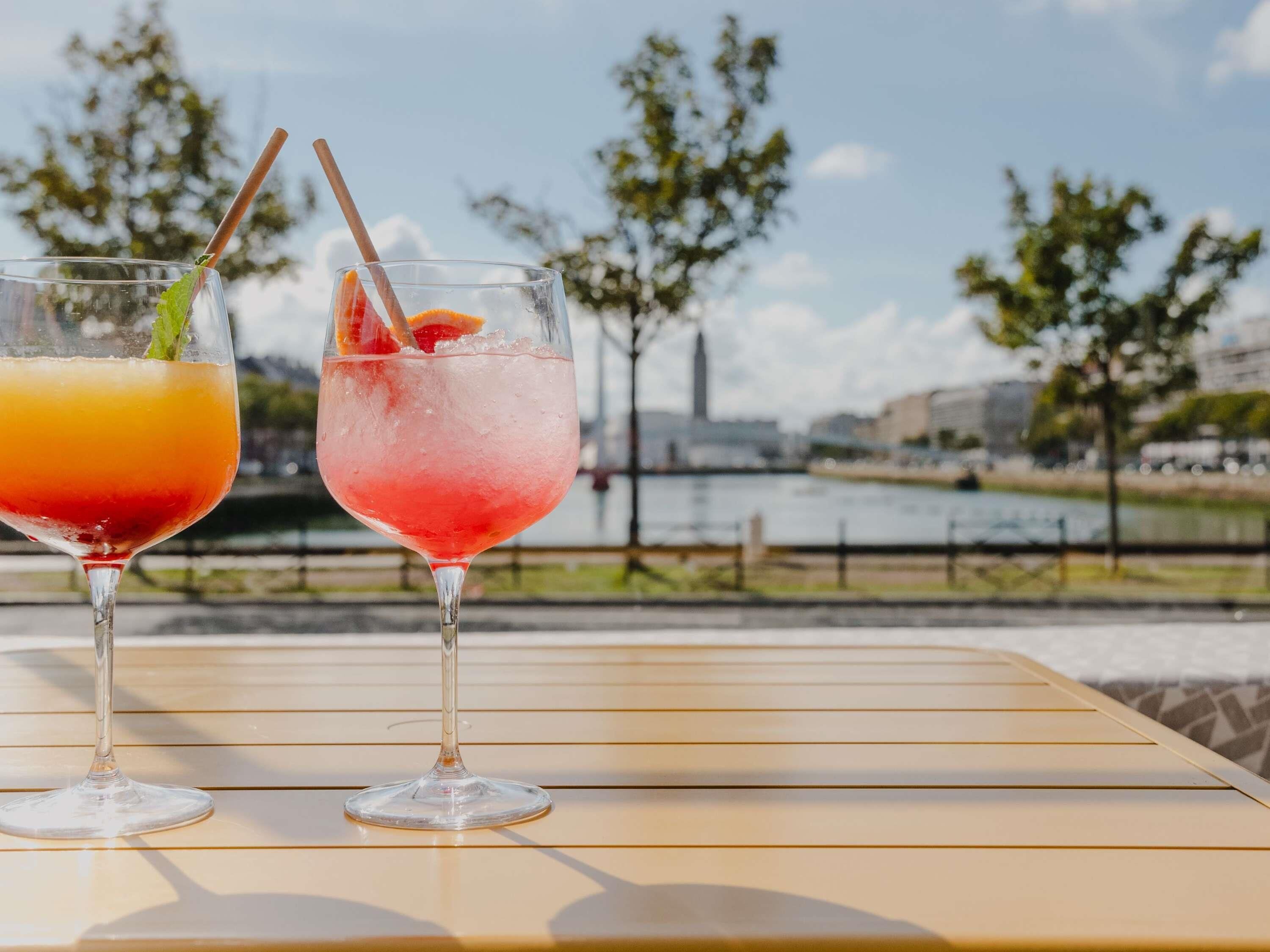 Two drinks on a table with a view of a river and buildings in the background.